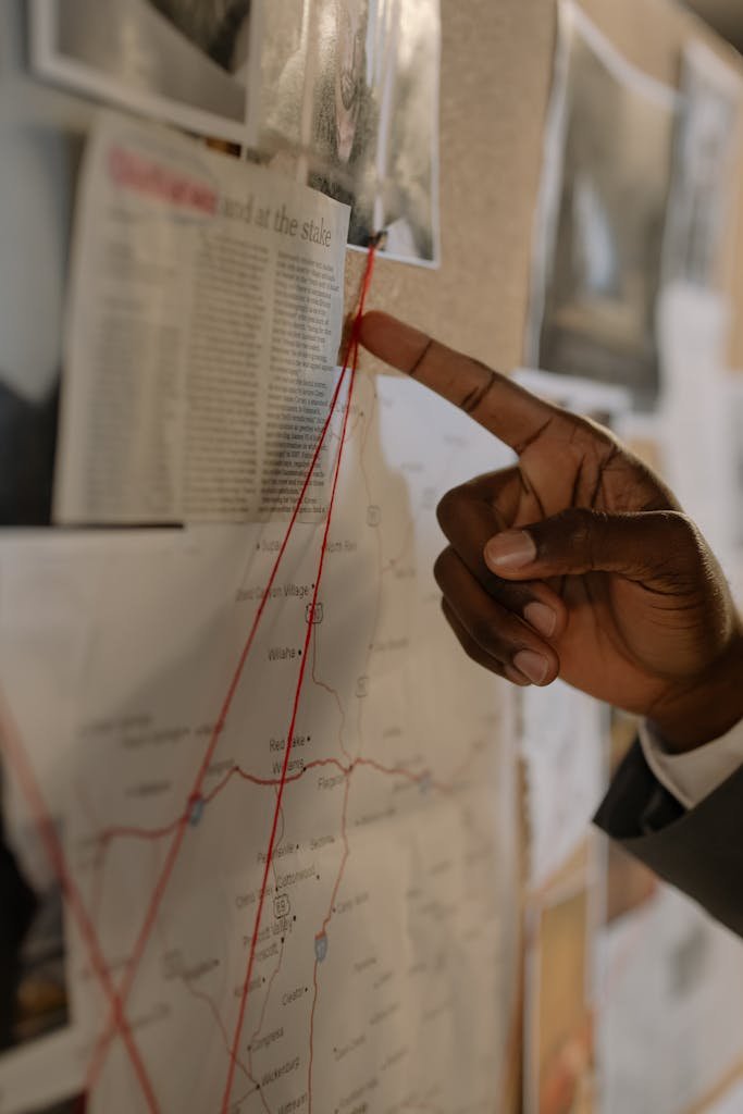 A hand pointing at a map with red strings on a corkboard, depicting an investigation setup.