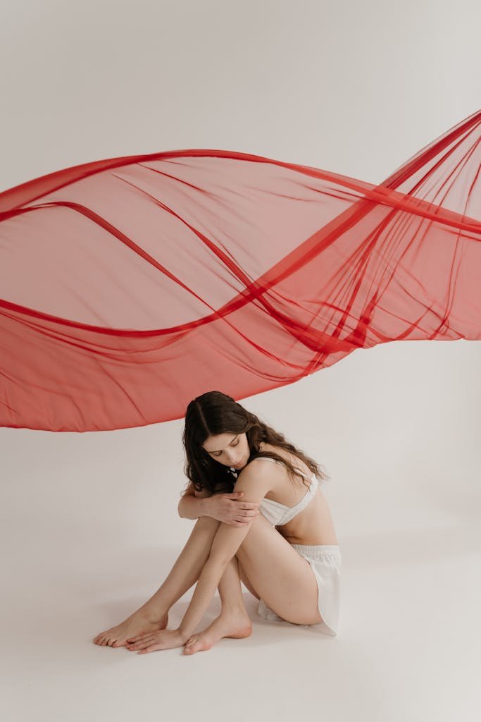 A woman sits gracefully beneath a swirling red fabric in a minimalist setting, exuding elegance.